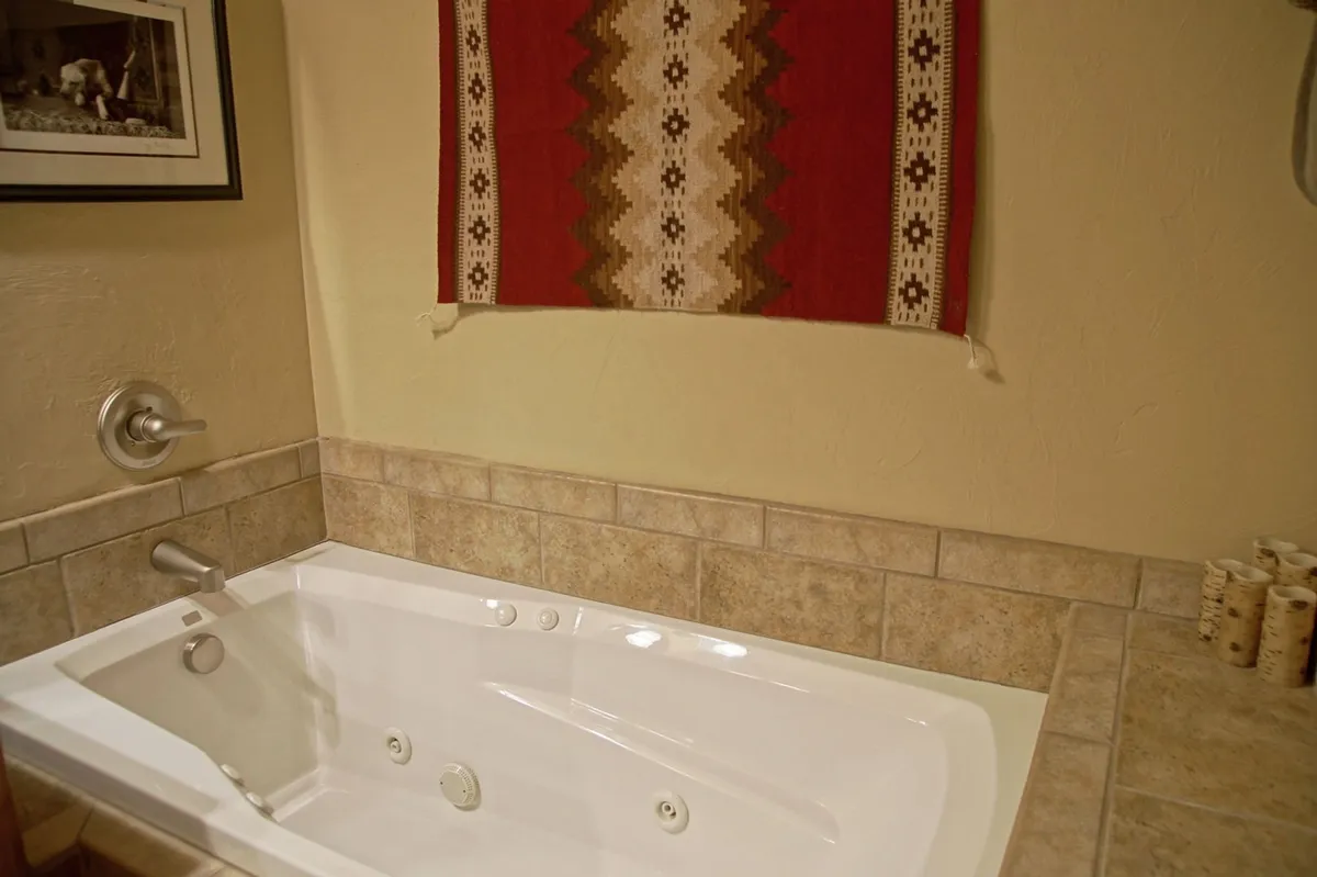A tranquil bathroom featuring a large whirlpool bathtub, woven wall hanging, and textured tile flooring. The setting is spa-like, offering a relaxing place to unwind after a long day.