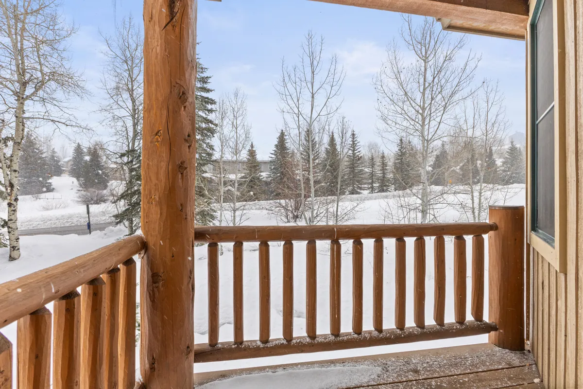 A tranquil balcony scene overlooking a pristine, snow-covered landscape with bare trees. The wooden railing is frosted with snow, suggesting peaceful mornings or evenings spent enjoying the mountain air.