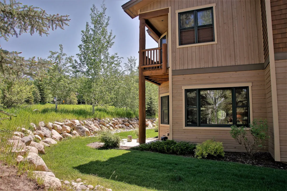 This image presents the townhome’s exterior, with striking wooden pillars, a stone pathway, lush greenery, and large windows. The inviting outdoor areas suggest a space perfect for gathering, relaxing in the garden, or enjoying the scenery. The setting reflects the summer atmosphere with well-maintained landscaping and outdoor-living appeal.