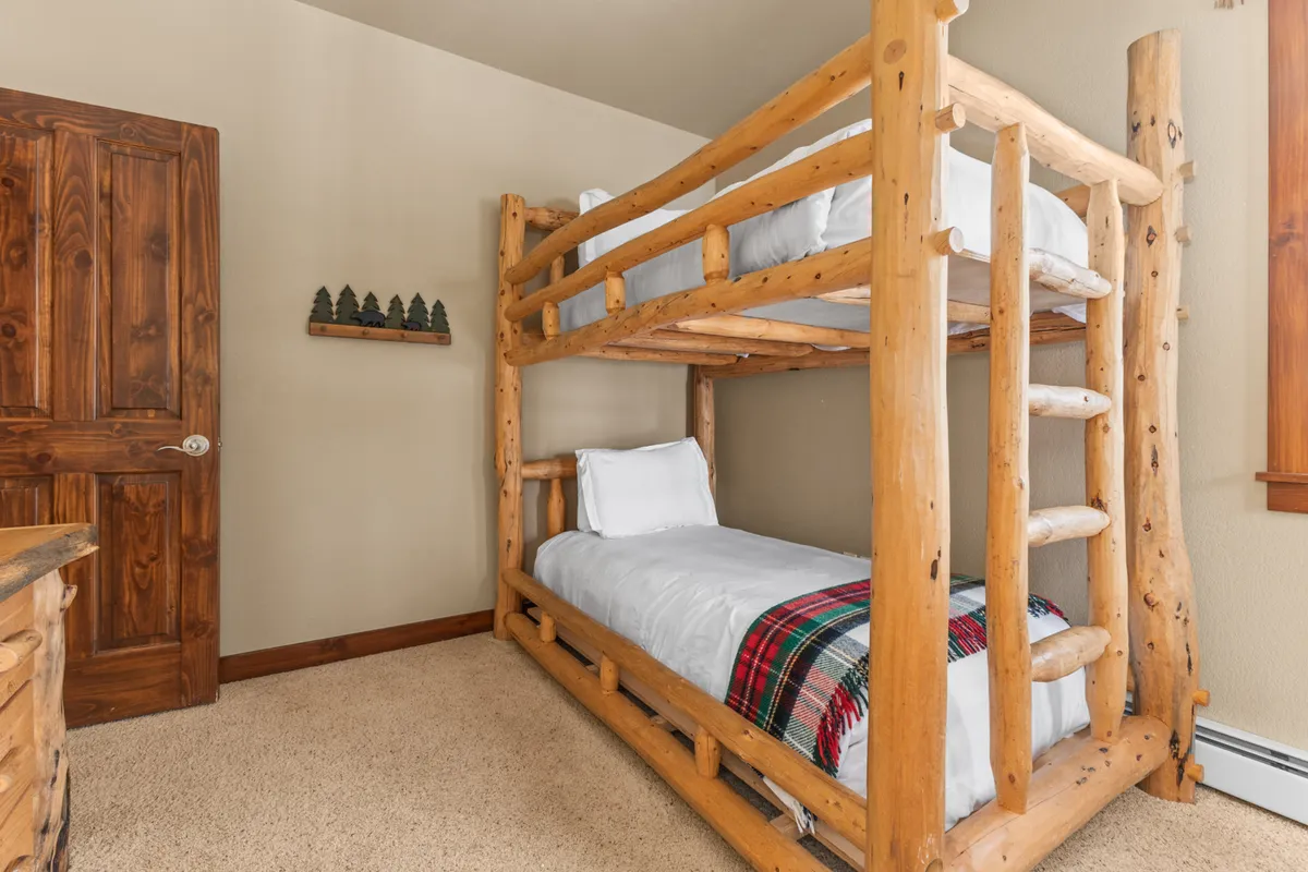 A warm, family-friendly bunk room with a rustic log-frame bunk bed, plaid blanket, wooden door, and a decorative shelf adorned with small tree figurines. The space feels welcoming and is ideal for children or friends, matching the description of the third bedroom.