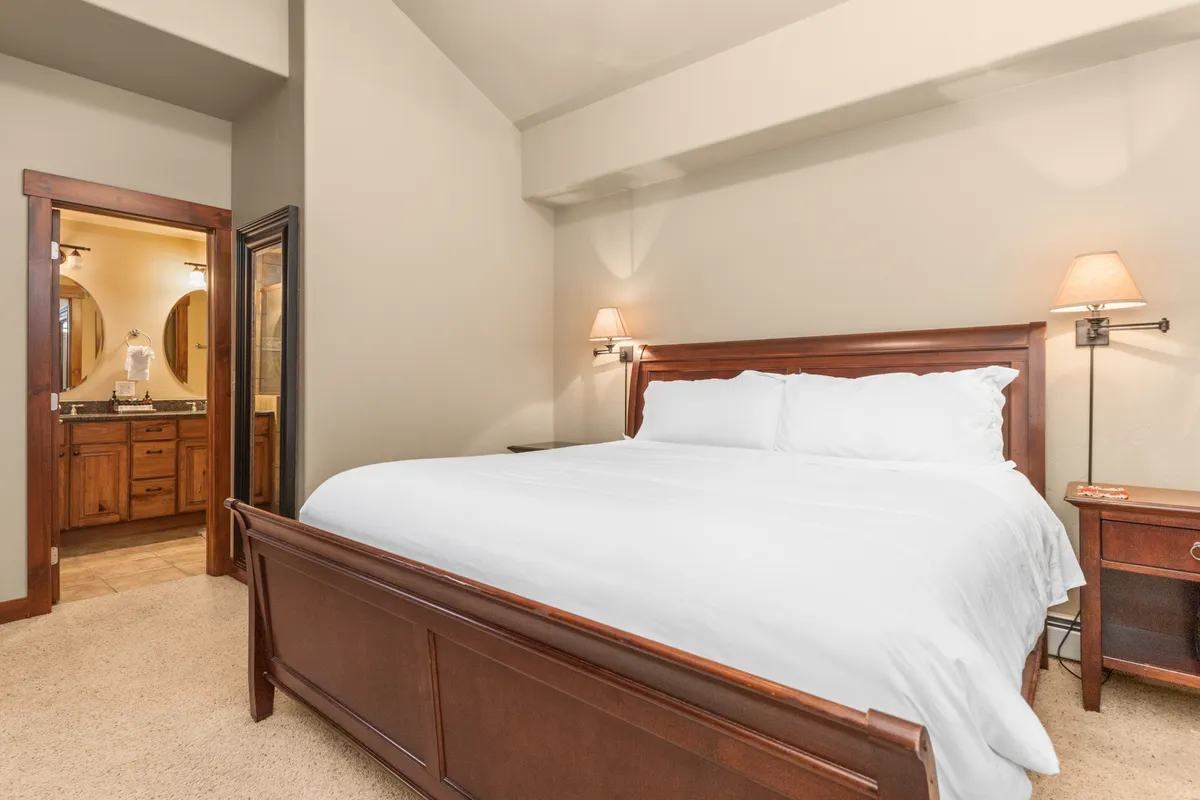 A serene king-sized bedroom featuring a plush bed with white linens, a nightstand topped with a lamp, and a cozy vanity area. The room exudes calm with its soft lighting and subtle decor, encapsulating the comfort and luxury of the primary suite.