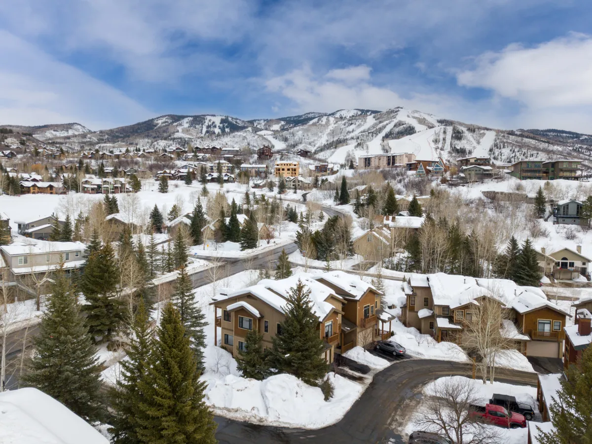 This exterior image captures snow-covered mountains rising behind rows of houses and pine trees, with a winding road leading toward the slopes. The landscape is blanketed in fresh snow, highlighting the proximity to ski and snowboard terrain.