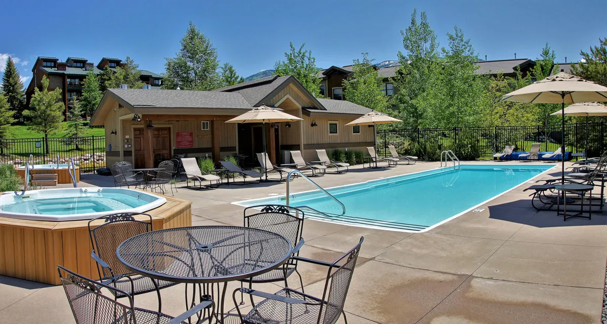 A scenic pool area featuring a swimming pool, adjacent hot tub, shaded outdoor seating, and umbrellas. The space is well-appointed for both socializing and quiet relaxation, with multiple seating options and lush surroundings.