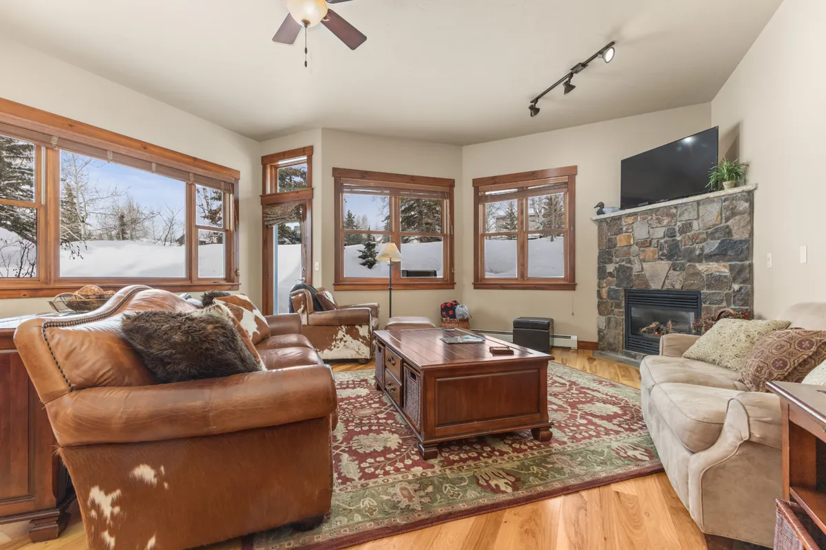 A spacious living room featuring a leather sofa set, a prominent stone fireplace, large windows offering plenty of natural light, and a wooden coffee table. The setting is warm and welcoming, ideal for communal activities or cozy gatherings.
