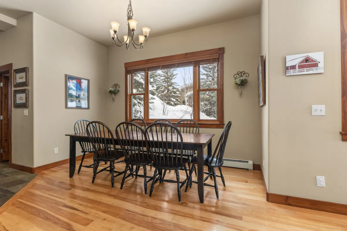 A welcoming dining area featuring a large table set beneath a striking chandelier, framed by natural light from nearby windows. The arrangement is designed for family dinners, festive gatherings, or casual meals, and flows organically from the open-plan living space.